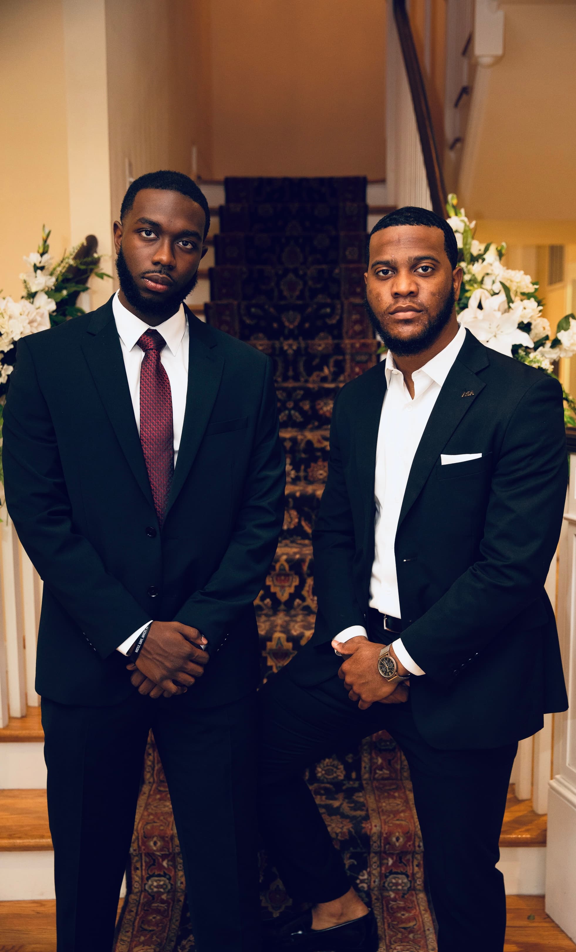 Mel and Mo in dark suits, standing side by side at the foot of a carpeted staircase lined with white flowers.